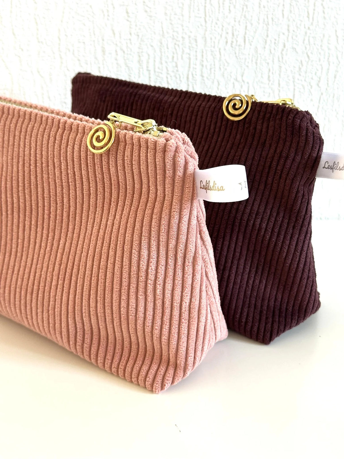 La pochette en velours côtelé rose blush devant la pochette prune.