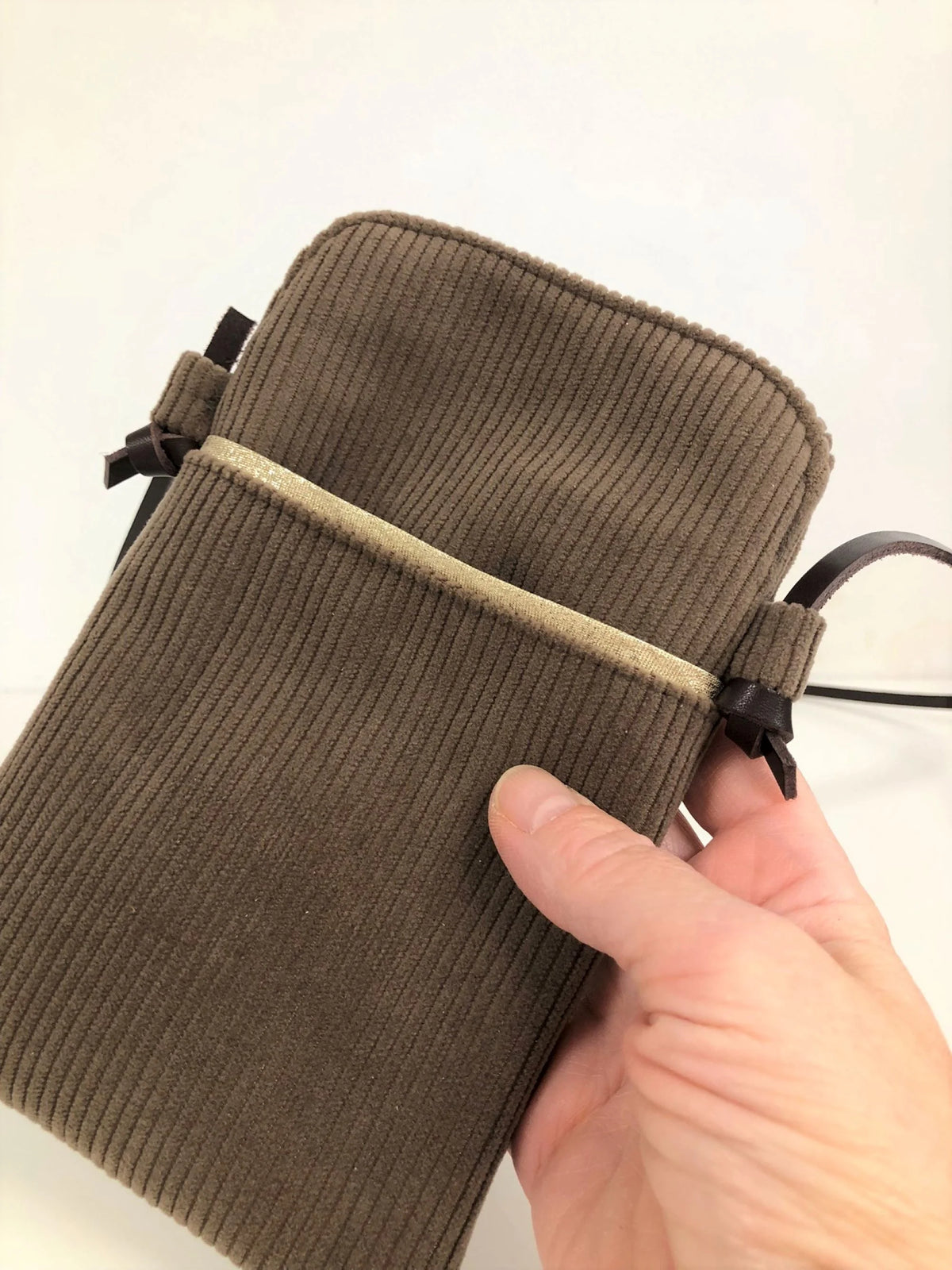 La pochette téléphone bandoulière en velours cotelé marron chamois et cuir, tenue en main.